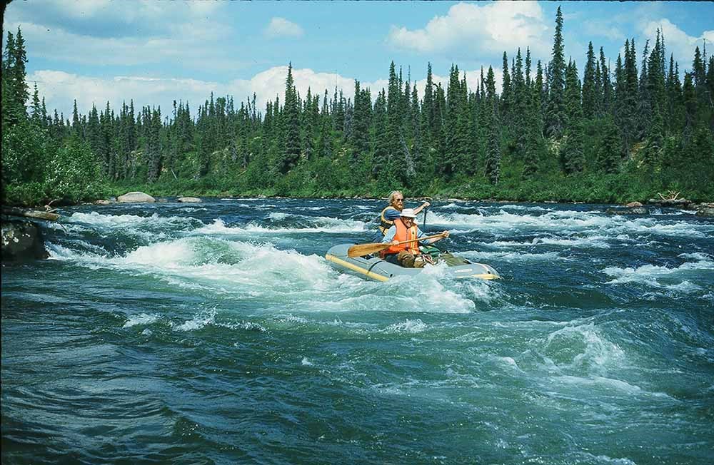 Two men in an inflatable raft float through rapids on a river lined with trees.