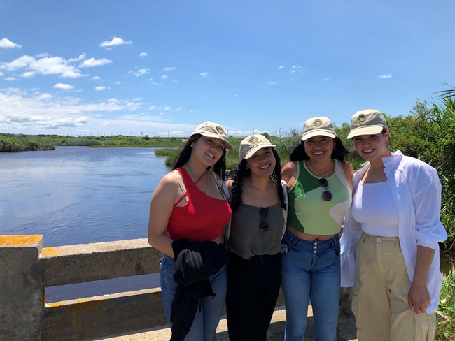 The four Mexican women stand on a bridge overlooking a salt marsh.