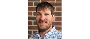 Man with brown hair, mustache, beard, and a white, blue and green plaid shirt smiling in front of a brick wall.