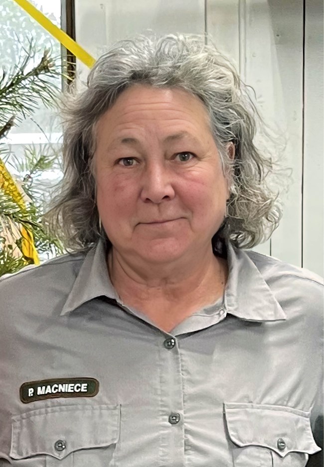Torso shot of woman in gray uniform shirt with shoulder-length curly gray hair in front of indoor plant