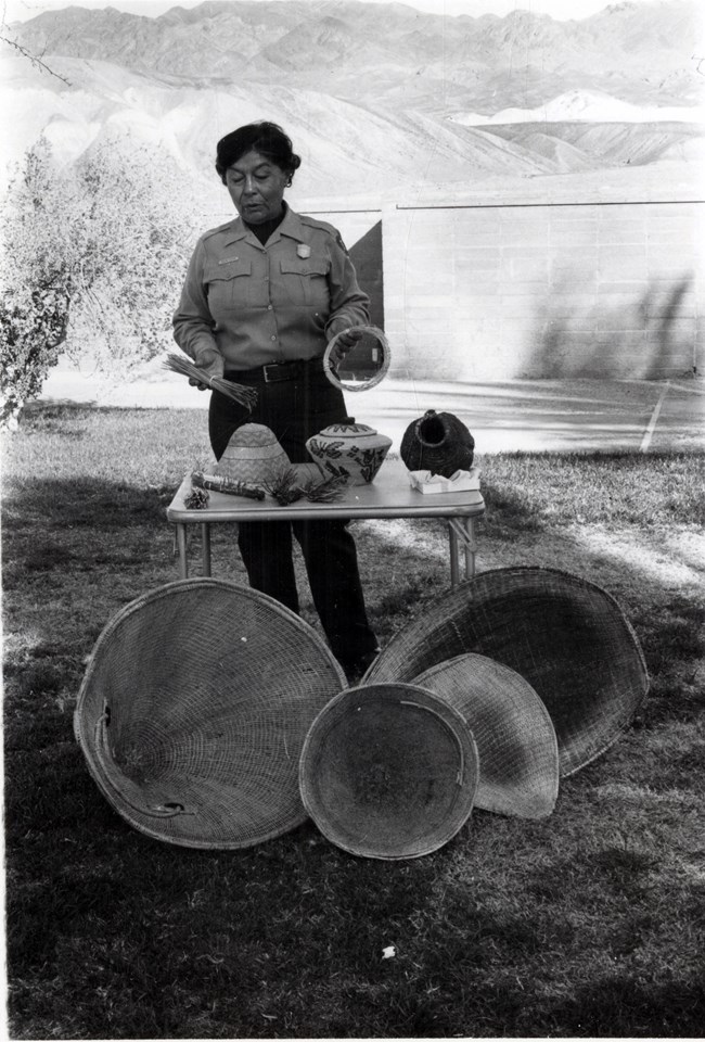 Pauline Esteves speaking in the Furnace Creek Visitor Center courtyard for the fifth anniversary of Death Valley National Park (1999). Native woman in a NPS park ranger uniform giving a talk. She is standing in front of a table with several native baskets.
