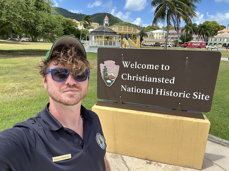 A young man wearing a hat and sunglasses in front of the Christiansted National Historic Site entrance sign