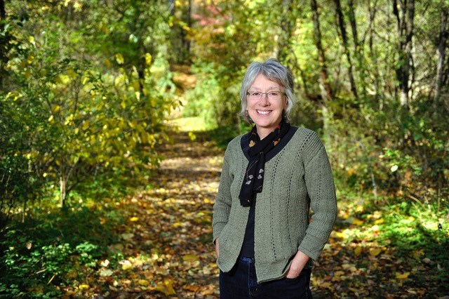 A smiling woman with grey hair and glasses stands in a trail scattered with fall leaves.