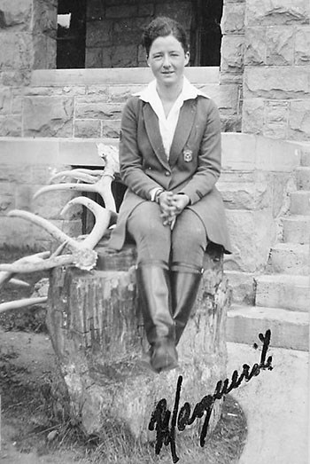 Marguerite Lindsley in her uniform with badge poses sitting on a stump of petrified wood with a large antler, in front of a stone building.
