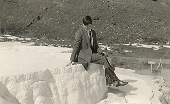 Marguerite Lindsley wearing a ranger uniform and holding a stetson hat sitting down on a natural feature with her ankles crossed.