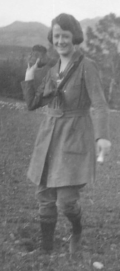 A woman wearing a long belted coat and knee pants with knee socks faces us with her right hand raised ready to toss something.