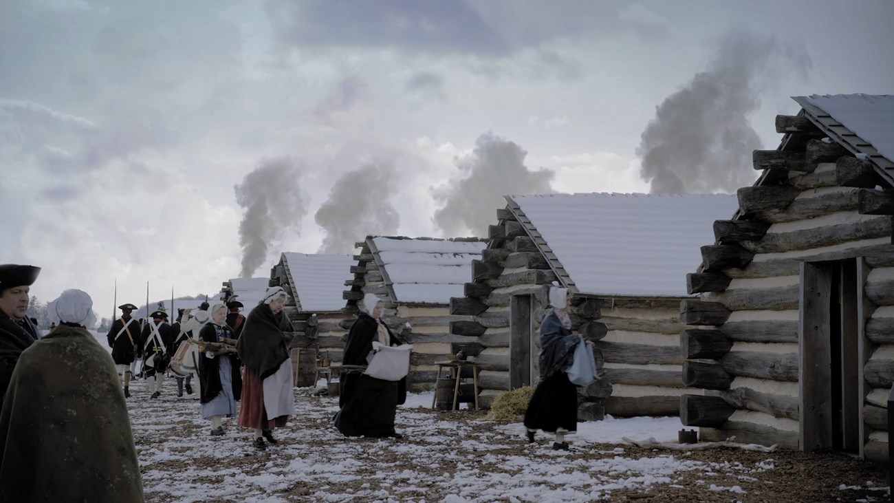 men and women in period clothing walk around log huts in the snow