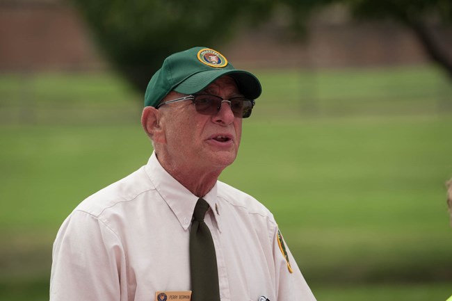 Perry talking about the flag to visitors. Elderly white male person, close up of his face as he speaks to other people. Background is grass with trees. He is wearing a white shirt with a black tie, and a green baseball cap that has the NPS volunteer logo.