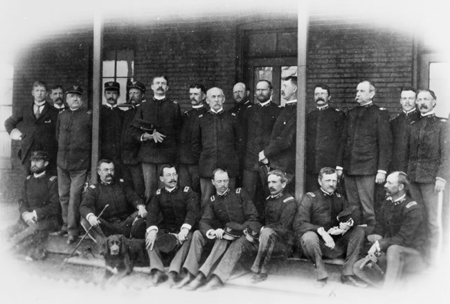 15 white men are standing and 7 white men are sitting in front of a brick building. They all wear the same uniform consisting of a dark jacket and gray pants