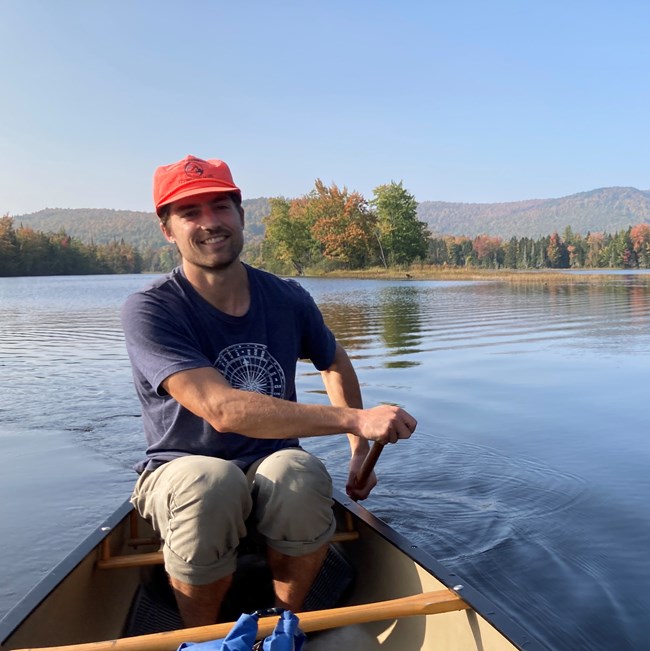 A man in a t shirt and orange hat canoing through calm water with rolling mountains and wetland behind him,