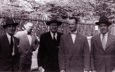 lt= Five men dressed in suits and ties standing in front of an iron fence.