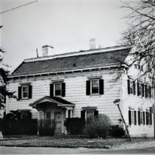 Large house, slanted roof, white siding, many windows, trees & bushes visible