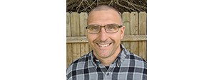 Man in black and gray plaid shirt and glasses smiles for a photo outside in front of a fence.