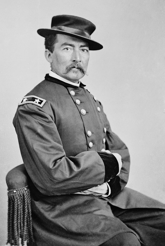 Black and white portrait of a civil war soldier in uniform wearing a hat, sitting on a chair, facing right