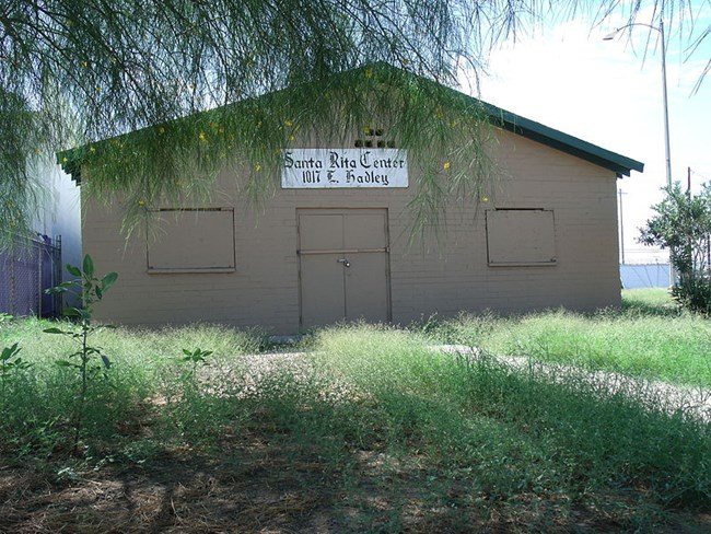 One floor brown building with a green roof. There is a tree covering the top left of it and grass surrounding it.