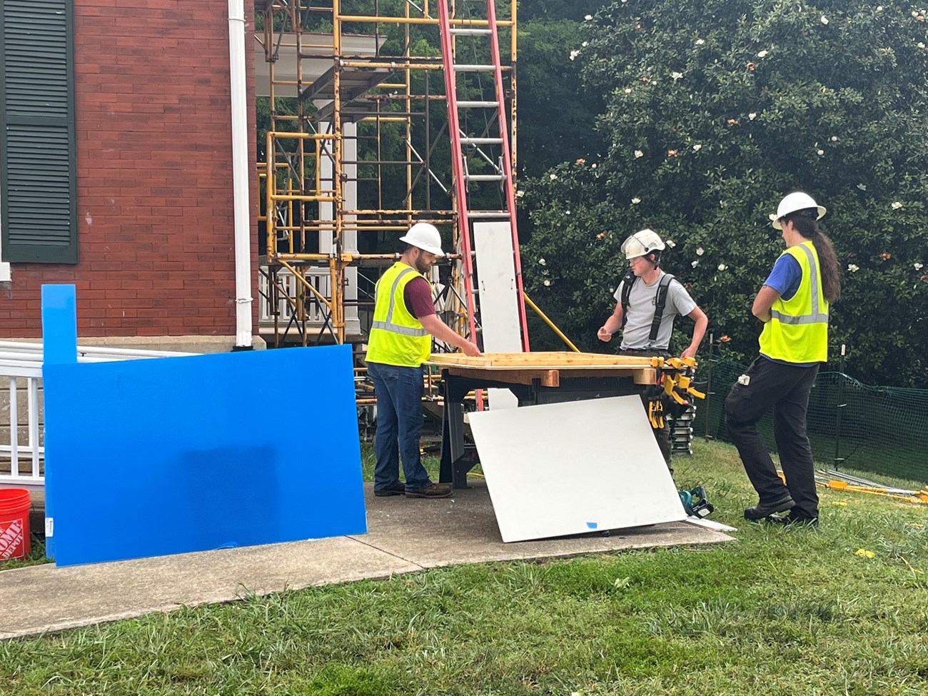 Three men handle a large square object below building scaffolding.=