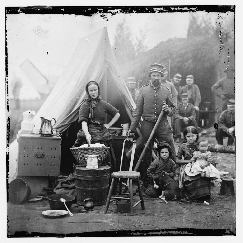 Tent life of the 31st Penn. Inf. The black and white photo depicts a civil war camp with a tent and uniformed men on bare ground. In the foreground, and woman with a tired expression and a hand on her hip holds a laundry basket. To the right of her is a uniformed man holding a saw.
