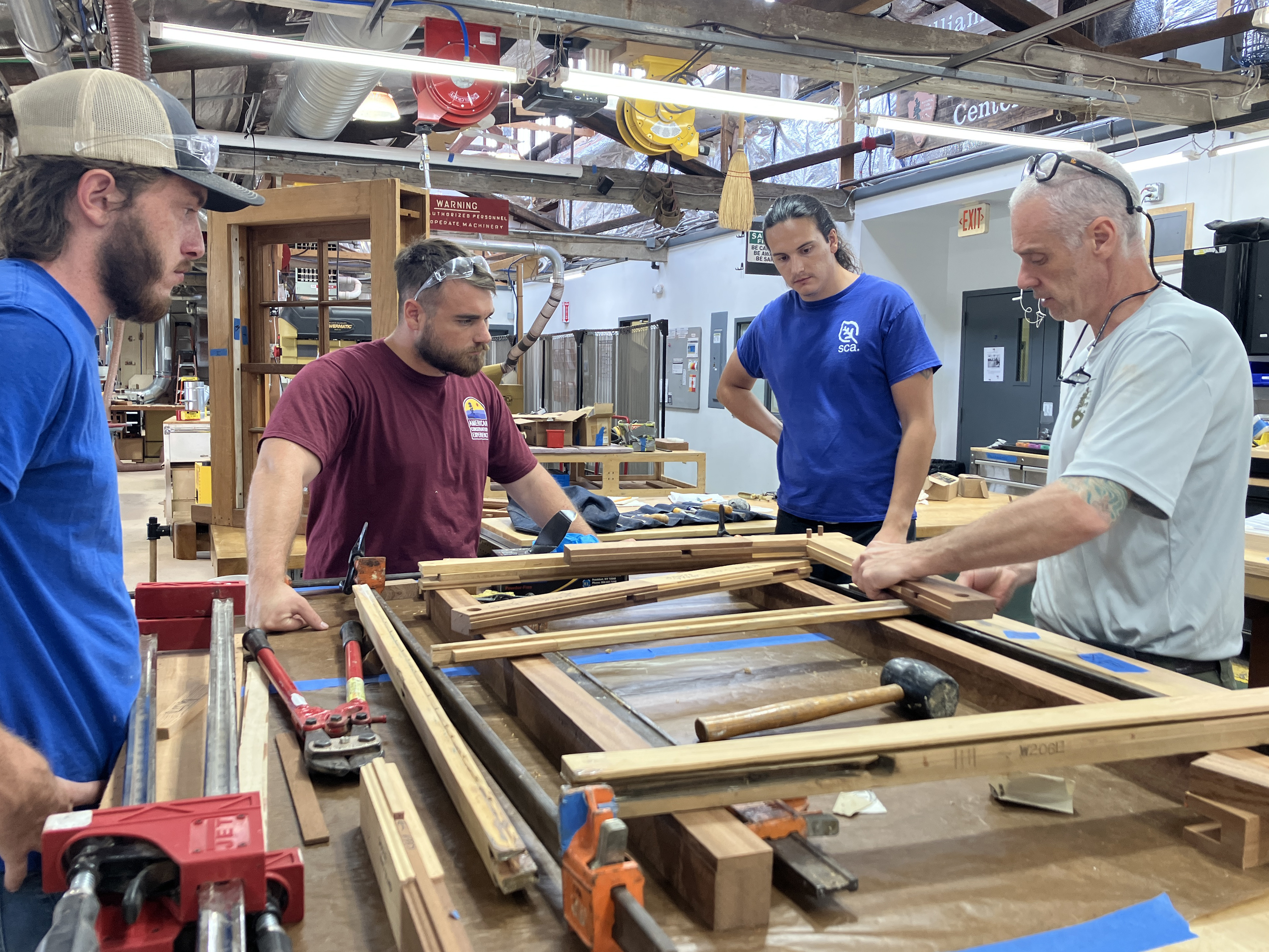 Three men watch an older man construct a window in a workshop.