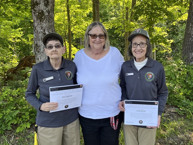 Two adults wearing volunteer polos and hats hold certificates, smiling next to a woman in a white t-shirt.