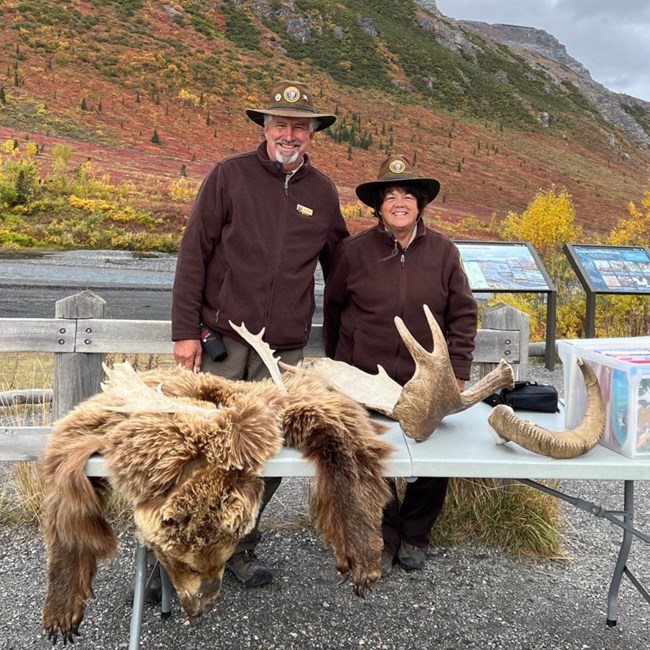 A man and woman wearing volunteer hats and jackets smile with a bear pelt and moose antlers in front of a mountainous range.