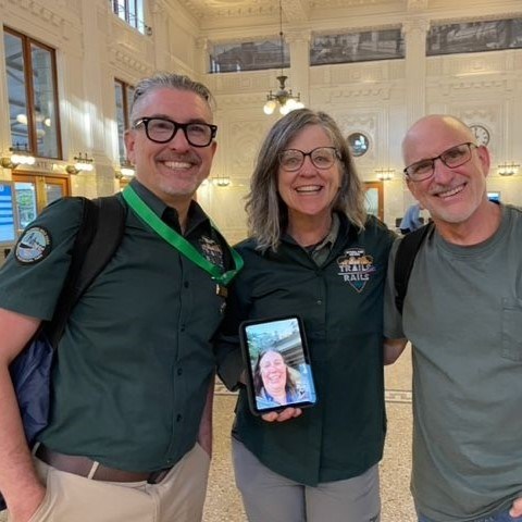 Two adults in Trails & Rails polos smile with another man and a woman on a tablet device.