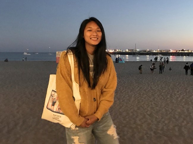Yvonne Ng Yvonne smiles at dusk on a beach with city lights behind.