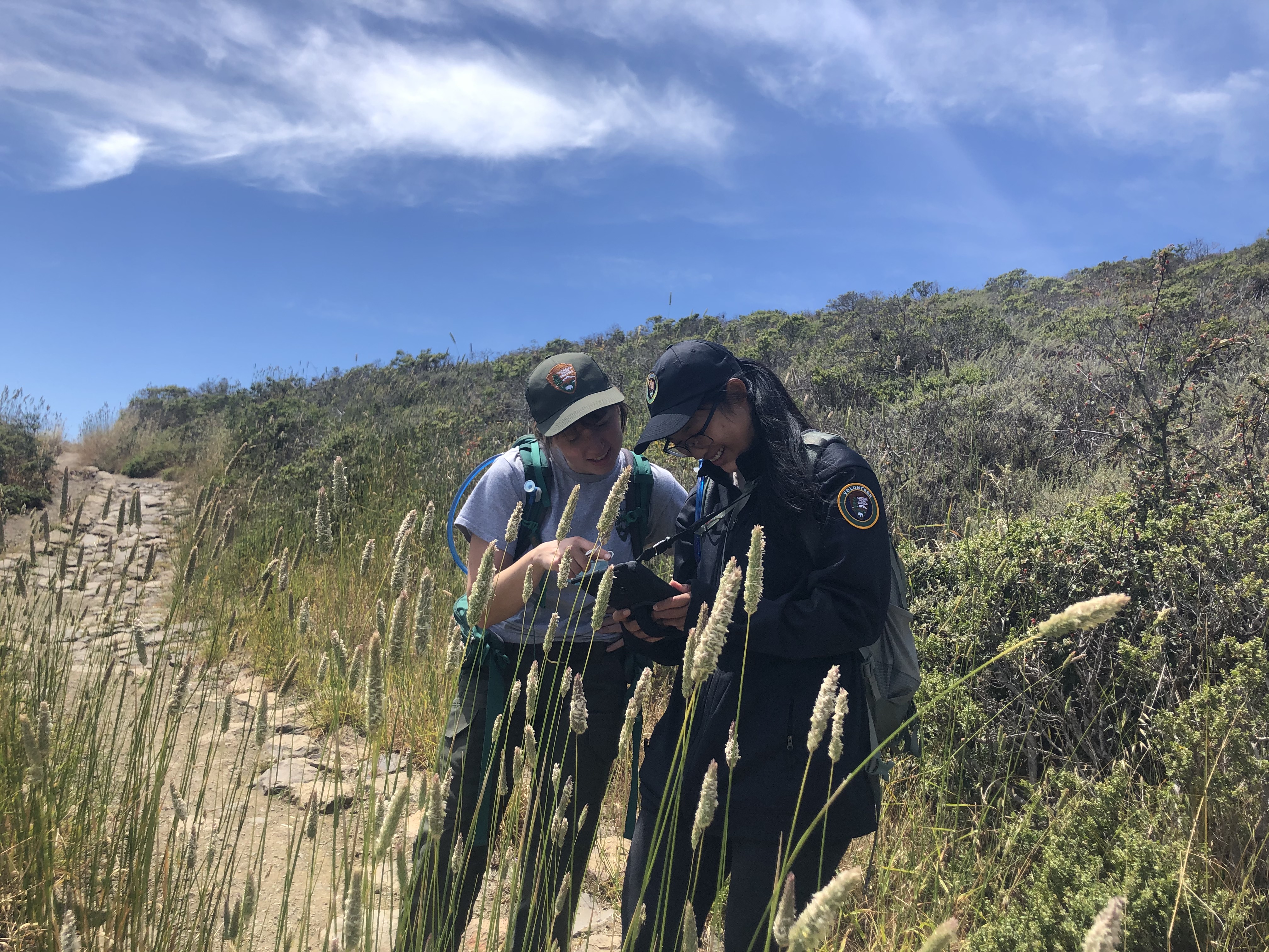 Two women looking at a tablet on a trail lined with tall grasses.
