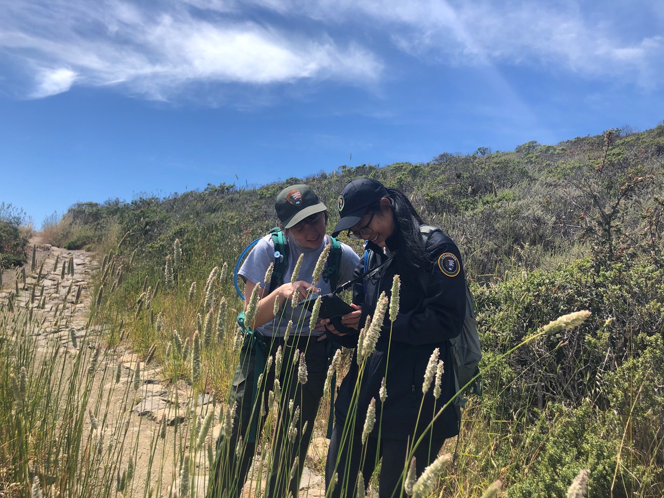 Two women looking at a tablet on a trail lined with tall grasses.