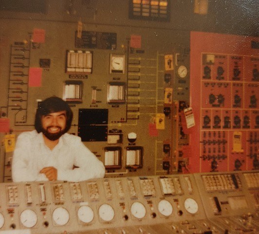 Carlos Leon Young man with a full beard and black hair stands between a bank of industrial gauges and in front of a wall with more gauges, dials, and controls.