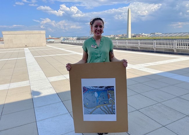 person holding an abstract drawing of a city, while standing in an open space and the Washington Monument in the background