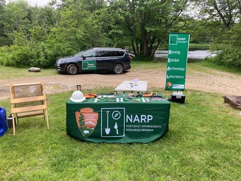 A table is set up with a table cloth that says NARP:Northeast Archeological Resources Program, it has a plethora of archeological tools. In the background is a sign that says Roving Archeological Resources Program and a Car with NARP on it.