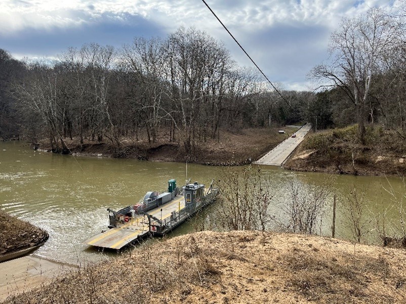 A vehicle ferry travels across a river toward a roadway with a car approaching the water’s edge.