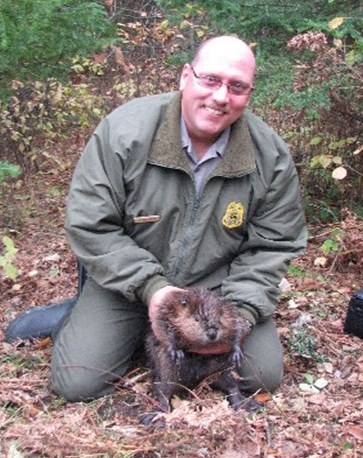 Park Ranger Kevin Grossheim. Photograph of person kneeling and holding beaver in the forest.