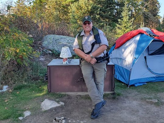 Park Ranger Kevin Grossheim Photograph of person standing next to tent and smiling at camera with trees in the background.