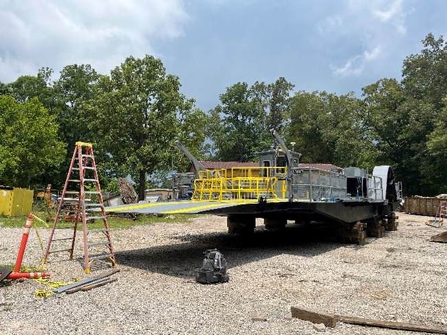 A ladder setup near a ferry boat drydocked in a gravel lot with green trees in the background.