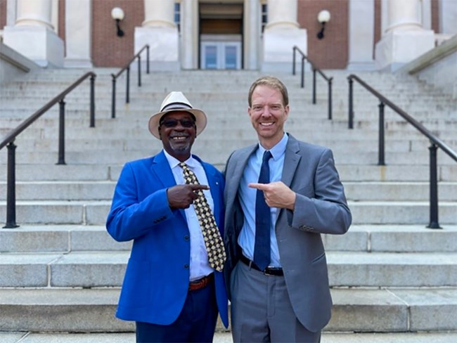 Two men stand at the base of a staircase pointing at each other and smiling.
