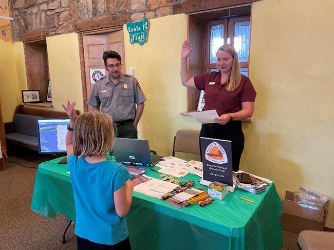Two park rangers stand behind a desk, holding up their hands as a child's back is to the camera with their hand up.