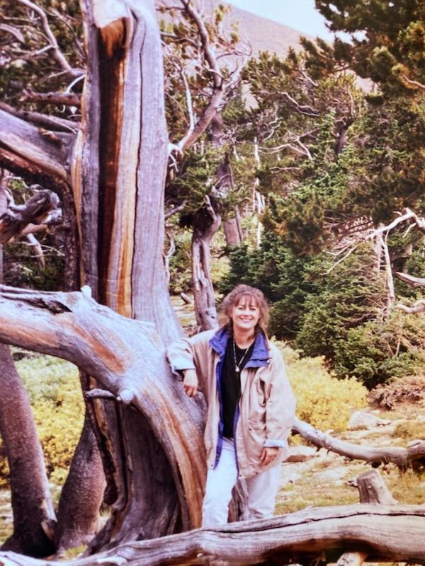 Smiling woman leans her arm against the trunk of a dead tree and is surrounded by evergreens with a mountain in the background.