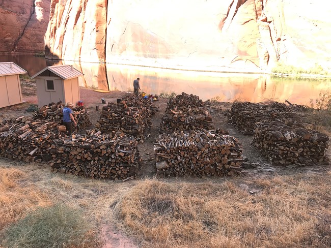 Piles of wood on the river shore. Two people are in he distance, and a couple of wooden bulidings.