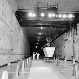 Pipe Gallery A black and white photo of a cavernous building. A pulley system is moving equipment through the building while two workers look on.