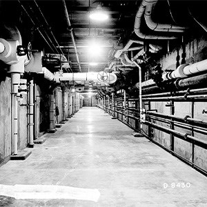 Pipe Gallery A black and white photo of a large concrete hallway with numerous pipes of varying sizes running along the walls and ceiling.