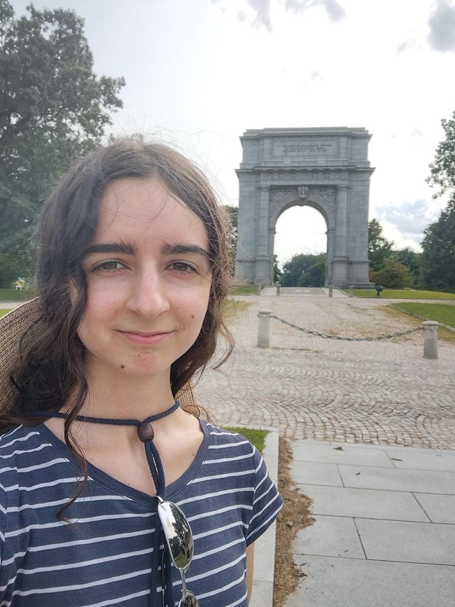 A person smiles in front of a large marble arch monument.