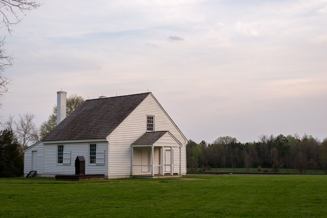 The image shows the small white wooden plantation office with a steep shingled roof, chimney, shuttered windows, and covered front porch of the building that Stonewall Jackson died in standing alone on a grassy field beneath an overcast evening sky, with