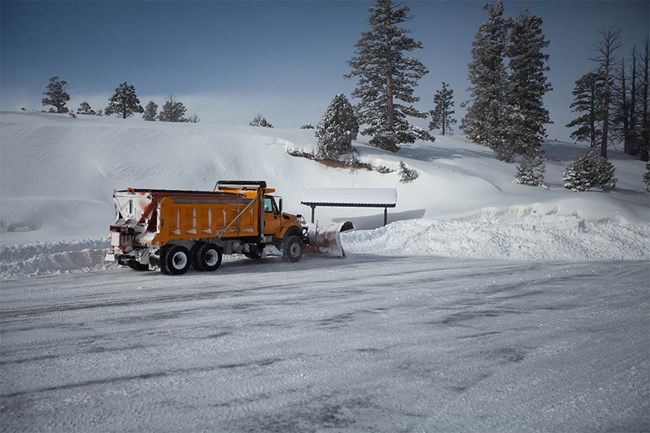 A yellow plow truck pushes snow from a large snowy parking lot