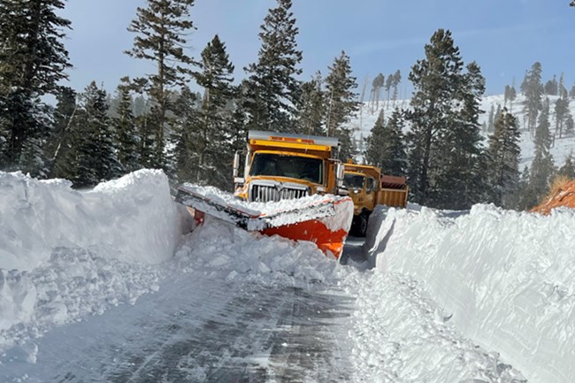 A large yellow plow truck pushes through multiple feet of snow along a road