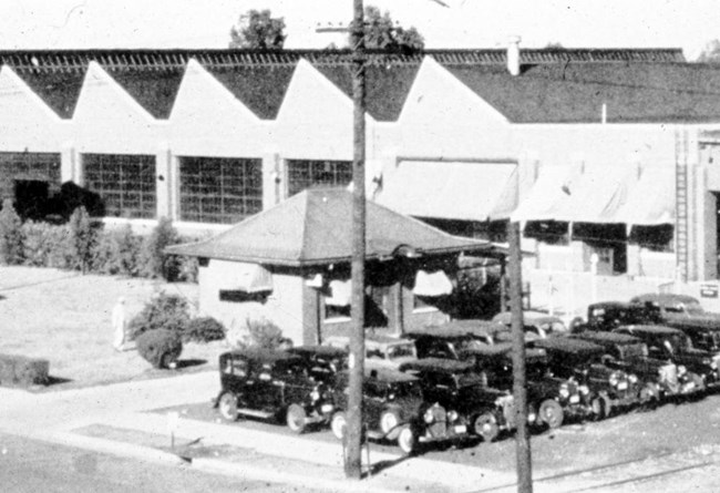 Two rows of cars parked in front of a small building with awnings and a hipped roof. The building sits outside the entrance of a large brick factory building with several sections of offset slanted roofs.