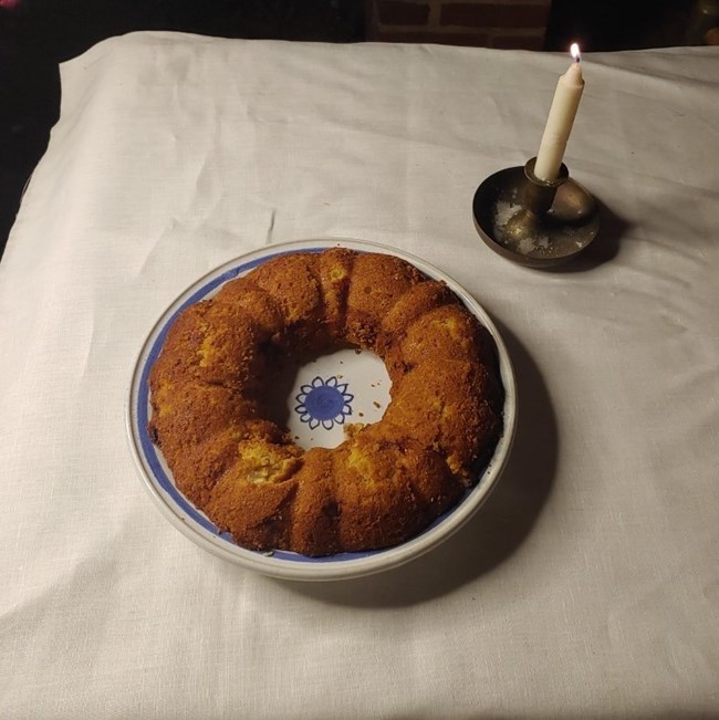 A bundt cake on a nice tablecloth. A single candle is lit next to it.