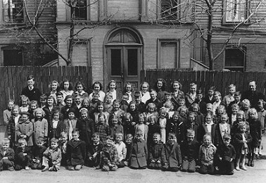 Black and white photo of children lined up outside the front of a wooden school building.