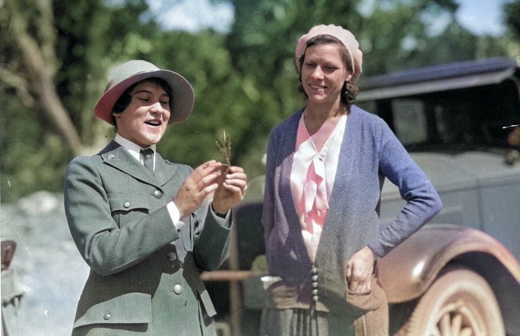 historic photo of a woman park ranger in uniform, talking with a visitor and describing the parts of a plan stem that she is holding