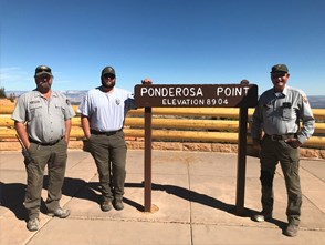 Three men stand together beside brown wooden sign reading Ponderosa Canyon